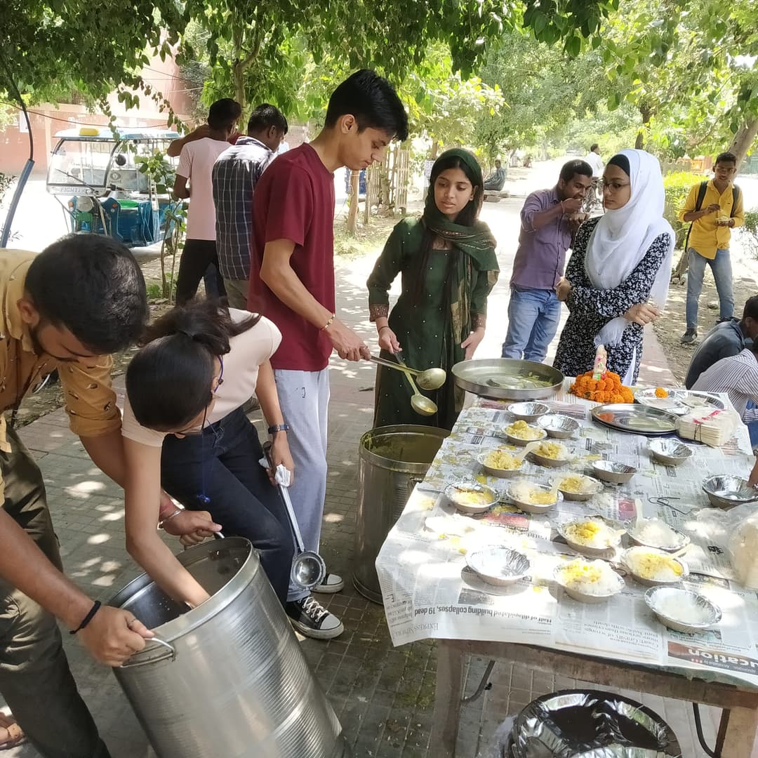 Helping Hut volunteers ladle freshly cooked food from large steel pots onto rows of plates laid out on a newspaper-covered table under the trees for community distribution.