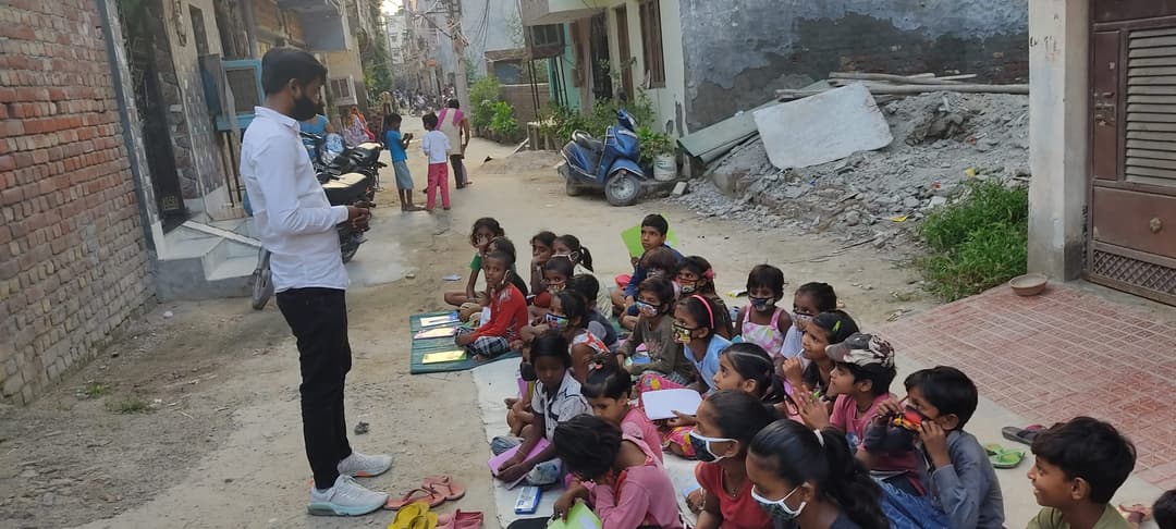 A Helping Hut volunteer stands in a narrow lane teaching rows of children seated on mats along the street during an Udaan Street School class.